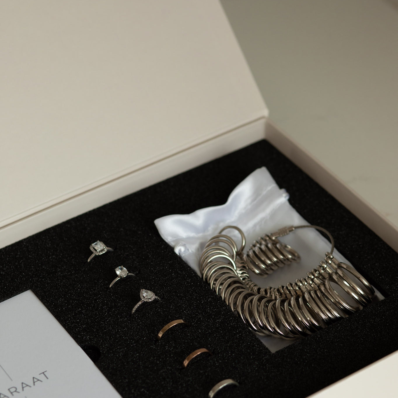 Jewelry sample box containing ring samples, a ring size gauge, and a white pouch, neatly arranged in a foam-lined case.