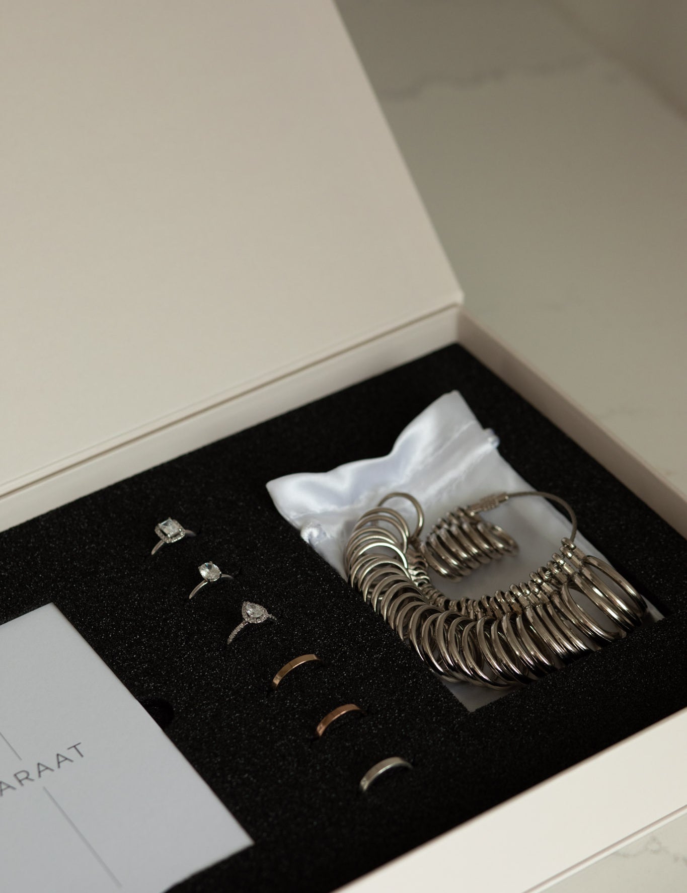 Jewelry sample box containing ring samples, a ring size gauge, and a white pouch, neatly arranged in a foam-lined case.