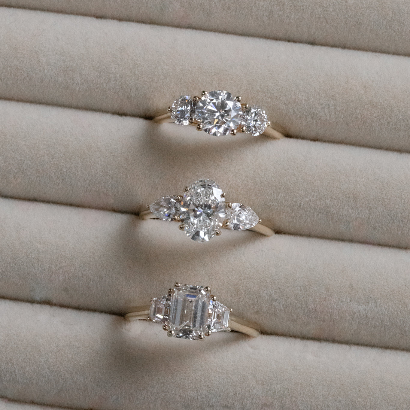 Three gold engagement rings with different diamond cuts (round, pear, emerald) displayed in a light velvet ring holder.
