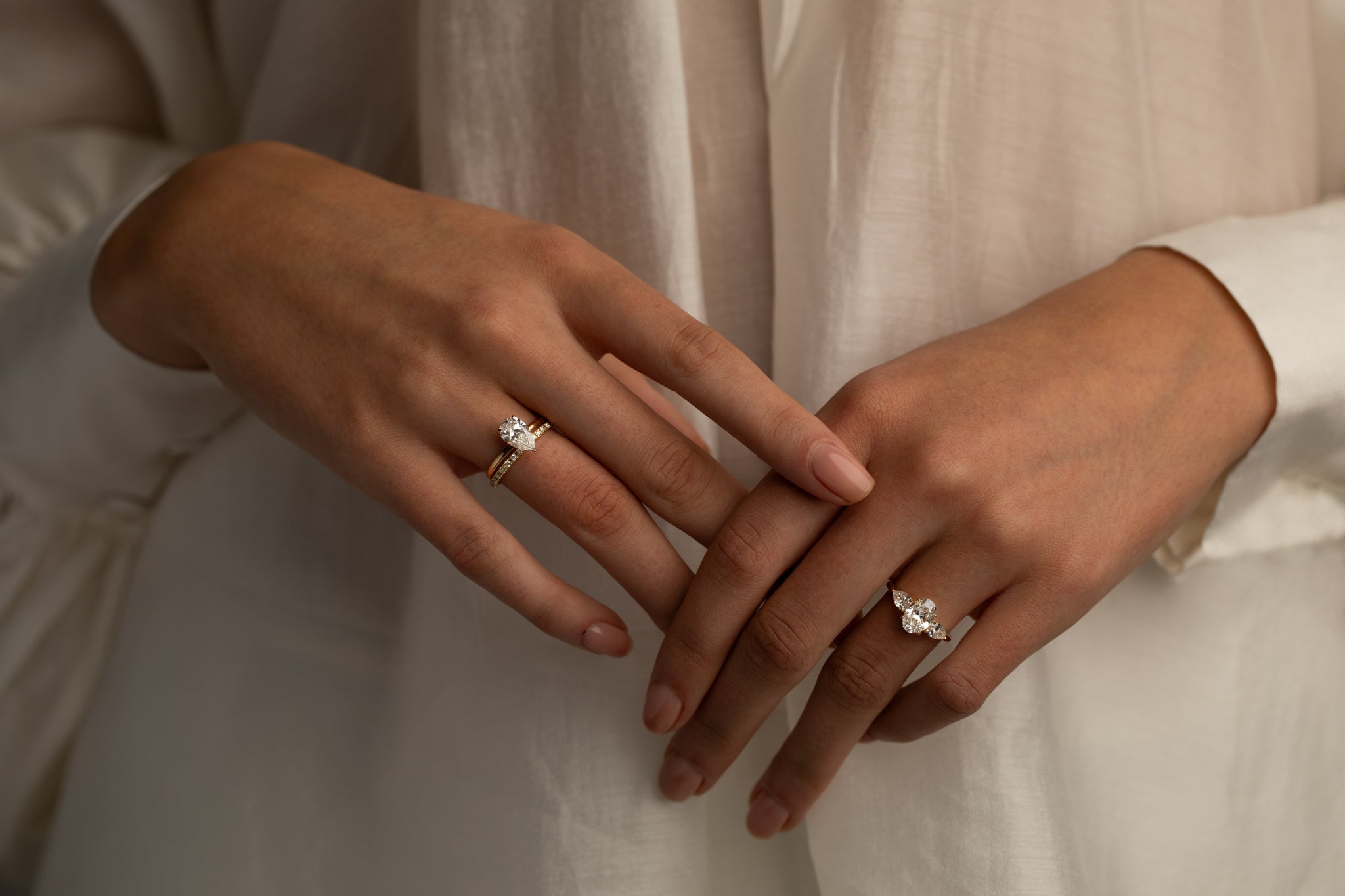 Close-up of two hands wearing elegant diamond rings with different settings, paired with a soft cream-colored outfit.