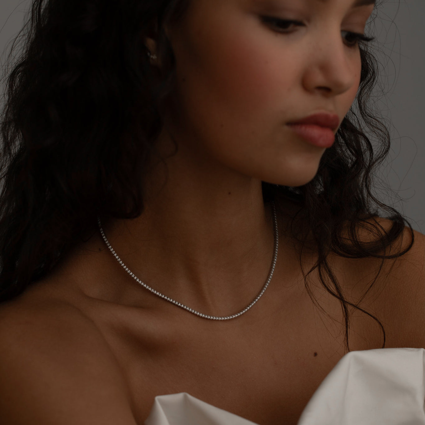 Woman with wavy dark hair wearing a delicate diamond tennis necklace, styled with a soft white fabric.