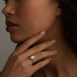 A close-up image of a woman's hand with a diamond ring on the finger, and small square diamond stud earrings. She is touching her chin while gazing slightly to the side.
