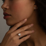 A close-up image of a woman's hand with a diamond ring on the finger, and small square diamond stud earrings. She is touching her chin while gazing slightly to the side.