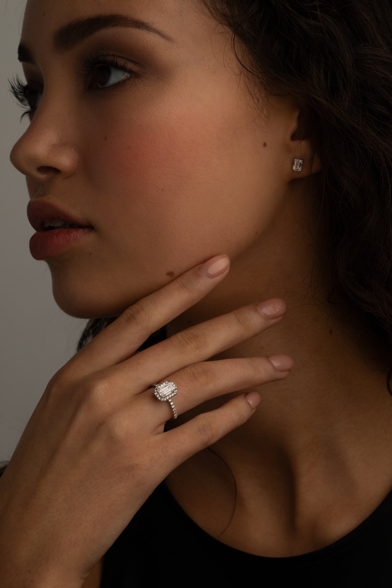 A close-up image of a woman's hand with a diamond ring on the finger, and small square diamond stud earrings. She is touching her chin while gazing slightly to the side.