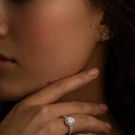A close-up image of a woman's hand with a diamond ring on the finger, and small square diamond stud earrings. She is touching her chin while gazing slightly to the side.