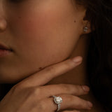 A close-up image of a woman's hand with a diamond ring on the finger, and small square diamond stud earrings. She is touching her chin while gazing slightly to the side.