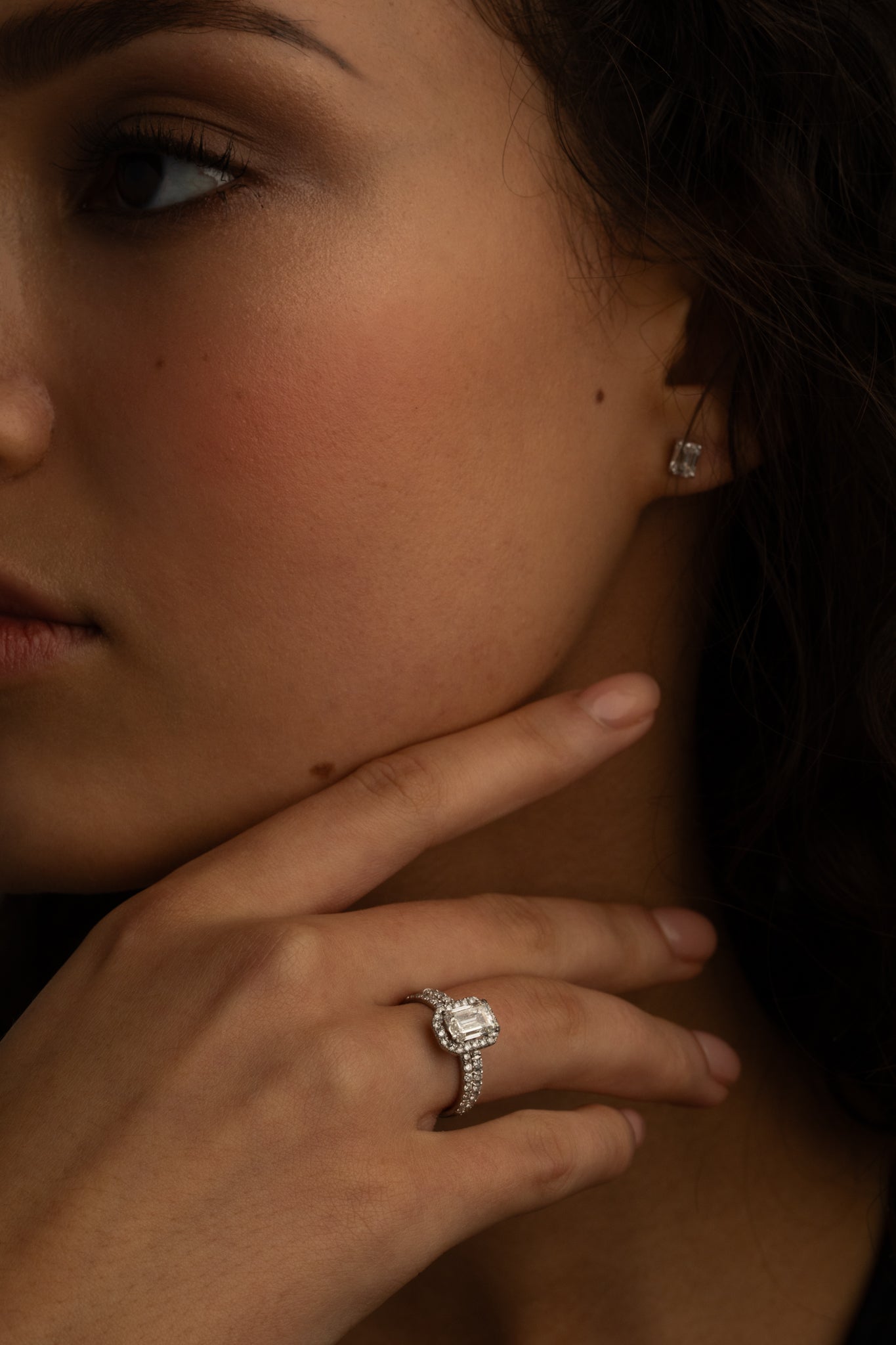 A close-up image of a woman's hand with a diamond ring on the finger, and small square diamond stud earrings. She is touching her chin while gazing slightly to the side.