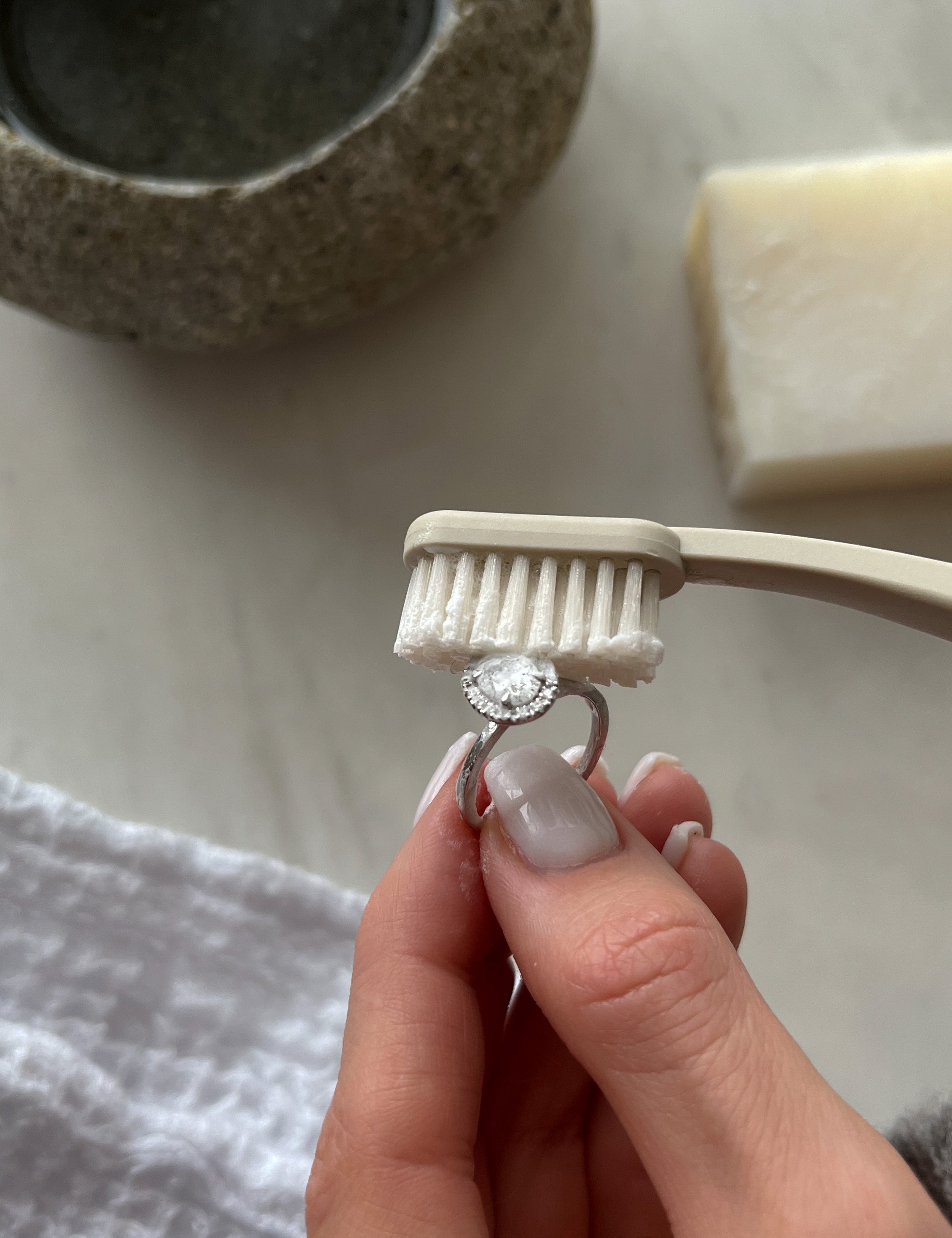 A hand holding a diamond ring being cleaned with a soft-bristle toothbrush, next to a stone bowl and a white soap bar on a marble surface.