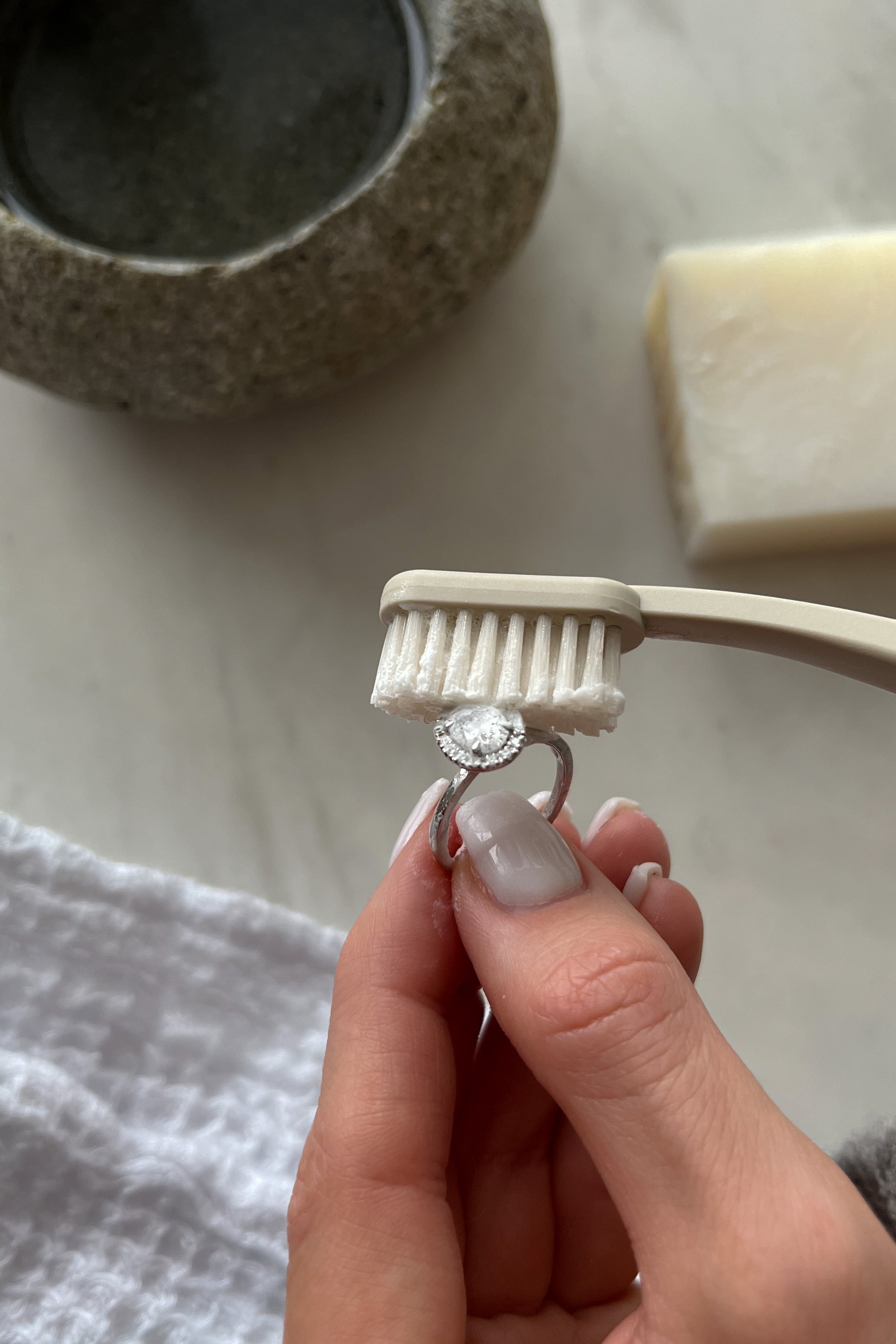 A hand holding a diamond ring being cleaned with a soft-bristle toothbrush, next to a stone bowl and a white soap bar on a marble surface.