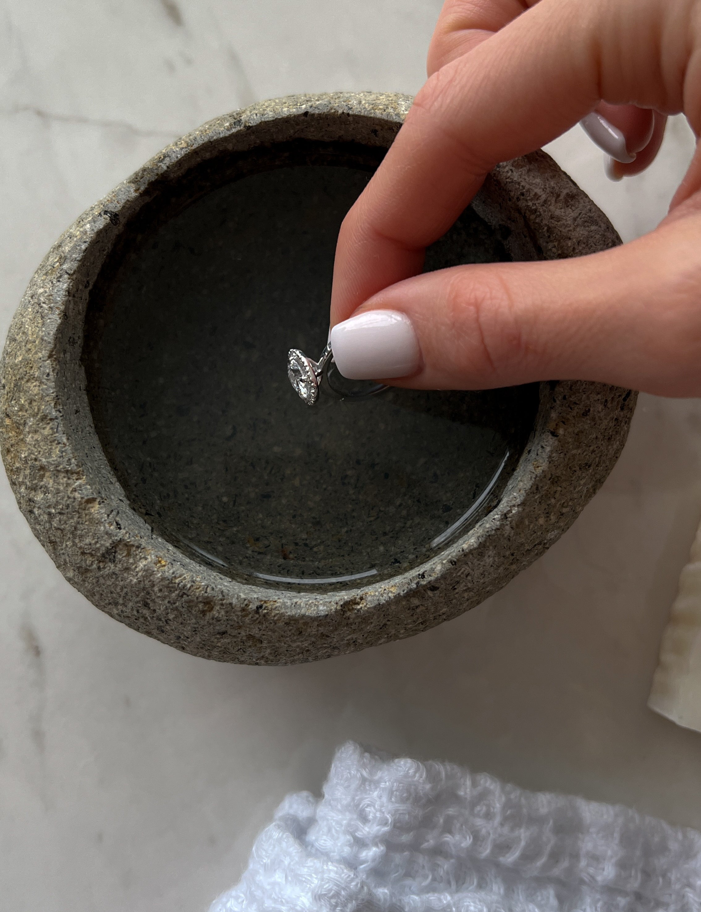 A hand with white-painted nails dipping a silver ring into a small round stone bowl filled with water, placed on a marble surface.