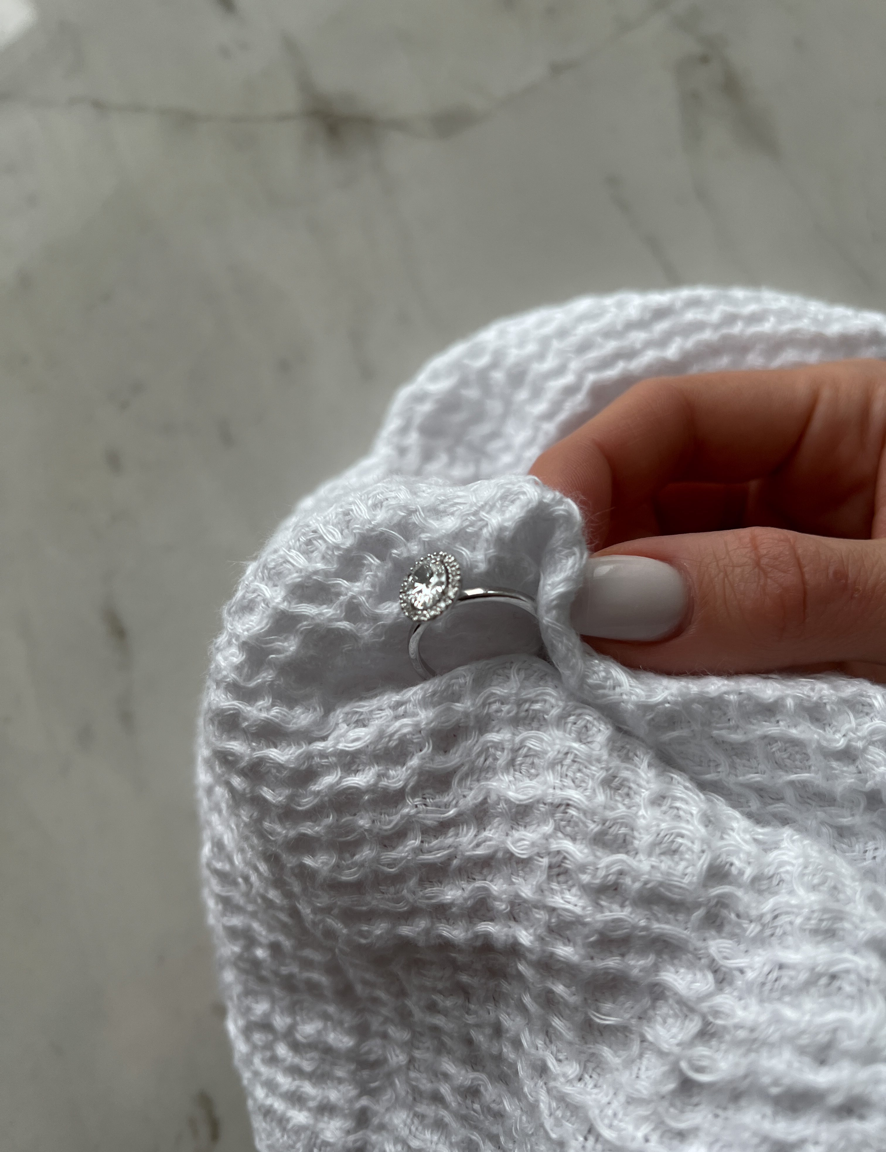 A hand with white-painted nails holding a silver ring with a diamond, wrapped in a white textured towel on a marble surface.