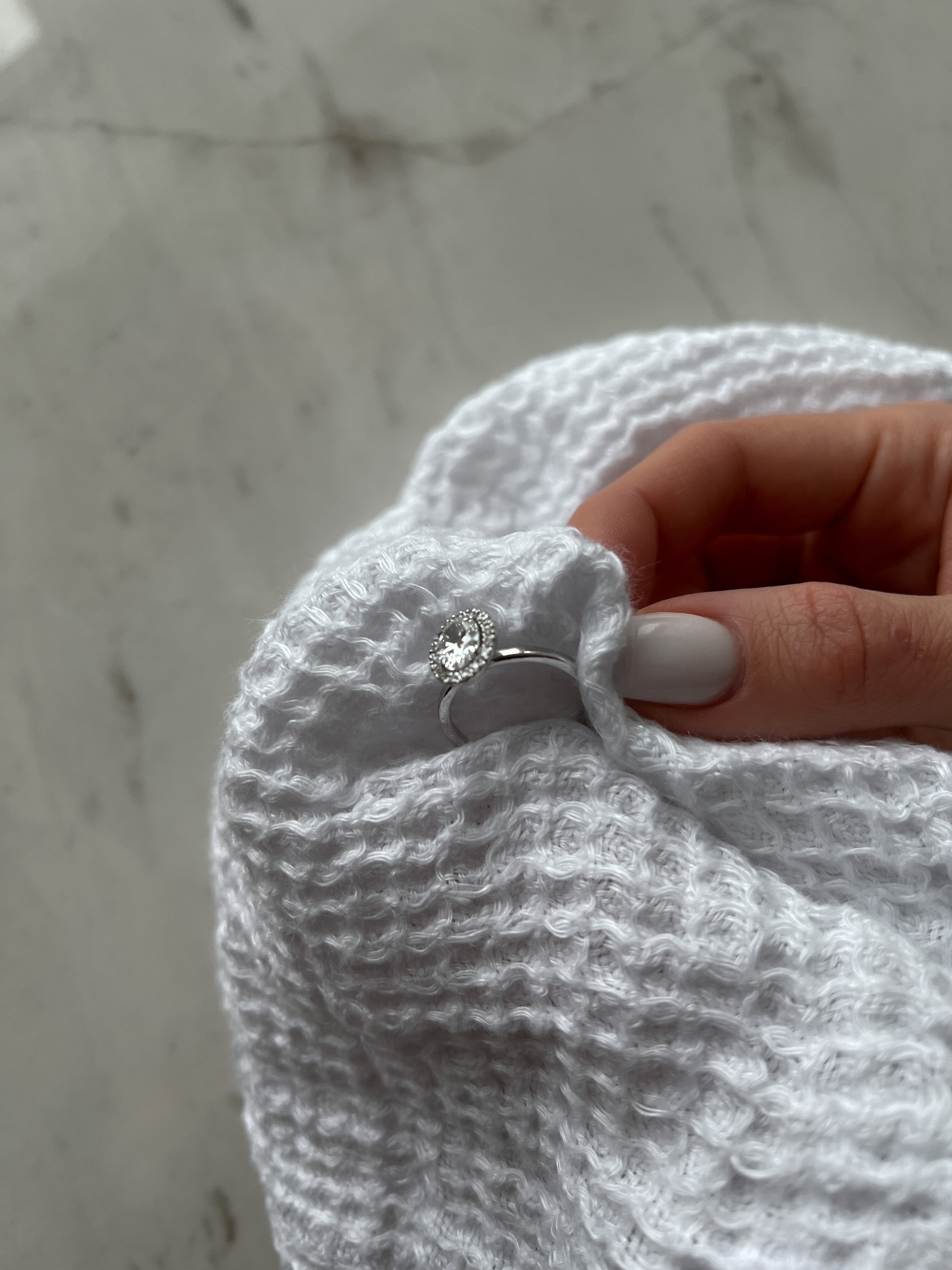 A hand with white-painted nails holding a silver ring with a diamond, wrapped in a white textured towel on a marble surface.