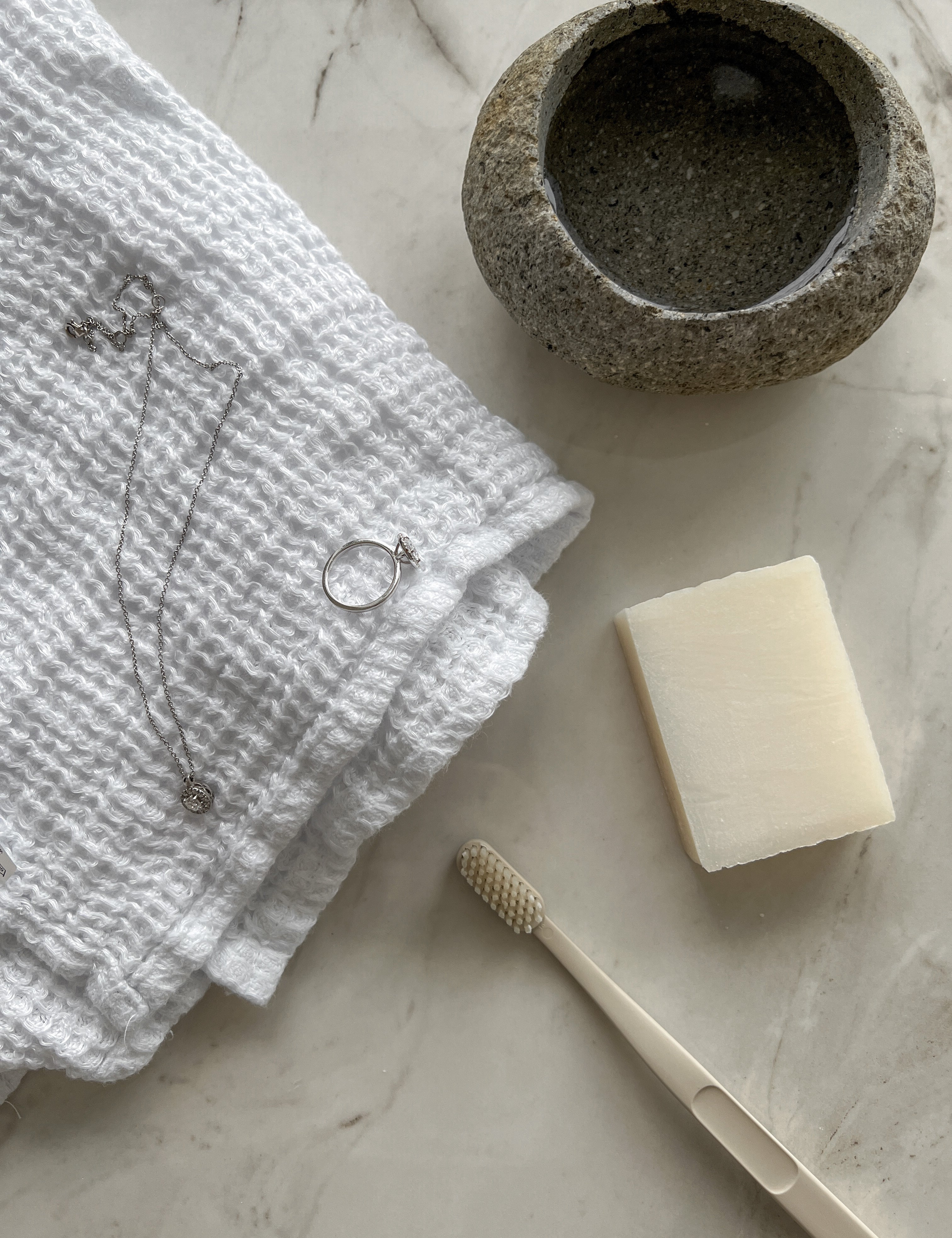 Jewelry cleaning setup featuring a white textured towel with a silver necklace and diamond ring, a stone bowl, soap, and a toothbrush on a marble surface.
