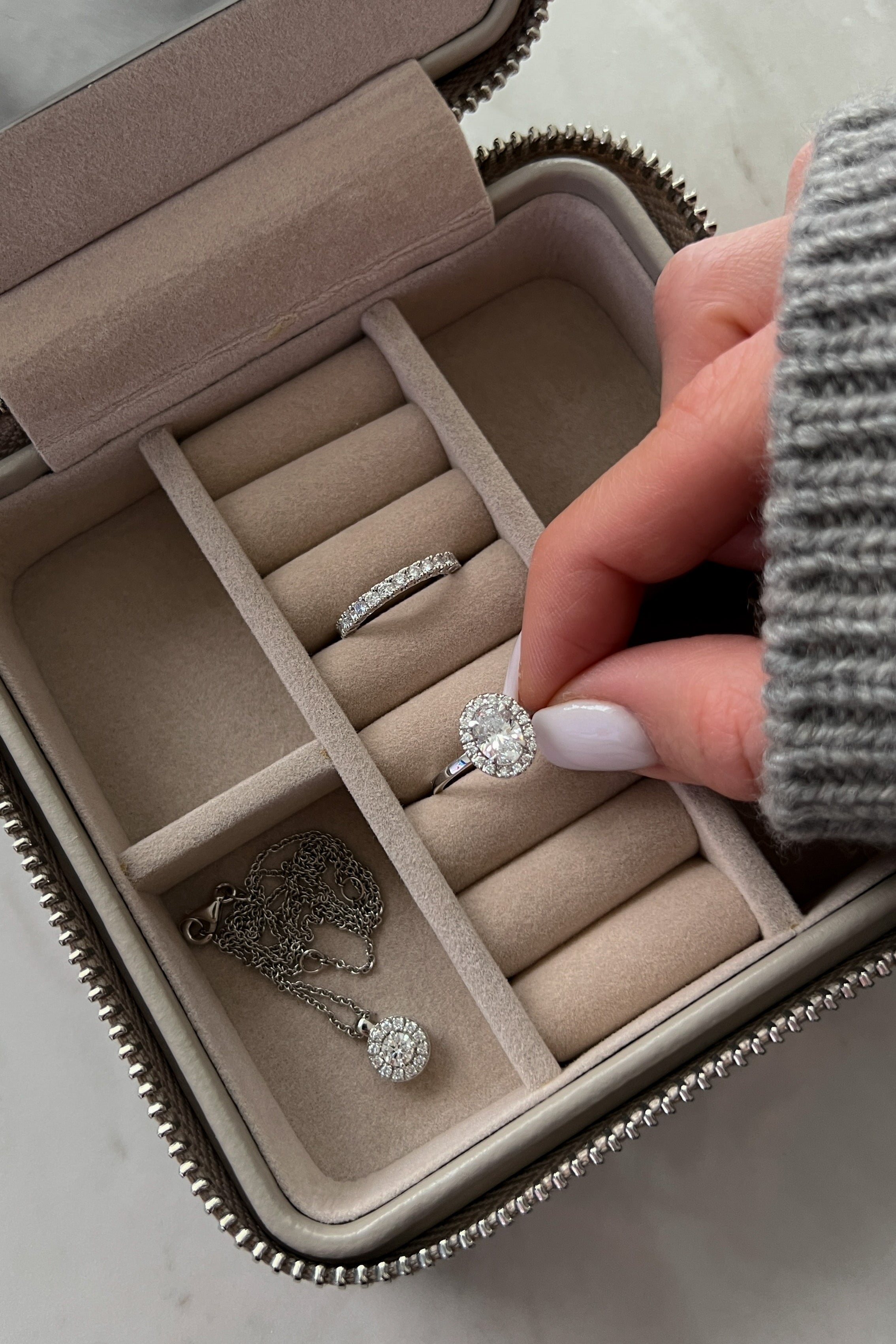 A hand with white-painted nails holding a diamond ring inside a beige jewelry box with compartments containing a necklace, ring, and band.