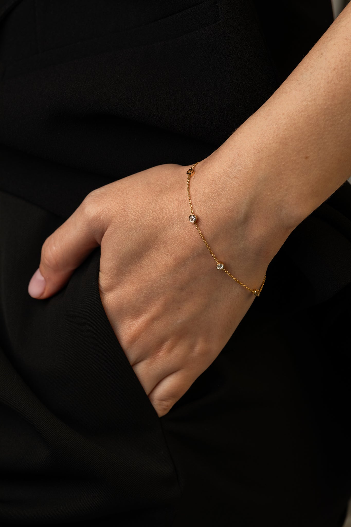 Close-up of a person wearing a delicate gold bracelet with small diamonds, resting their hand on the pocket of black clothing.