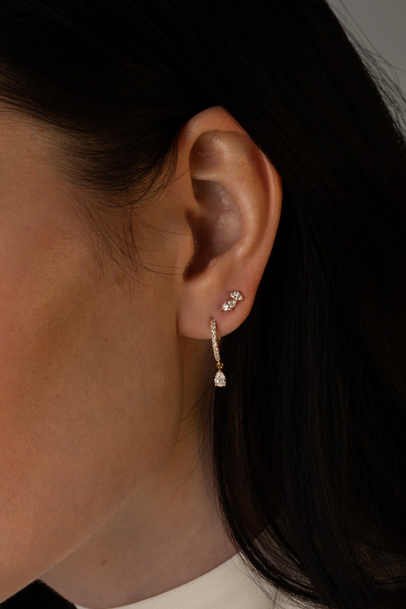 Close-up of a woman's ear wearing a small diamond stud earring and a gold drop earring with a pear-shaped diamond, paired with dark hair.