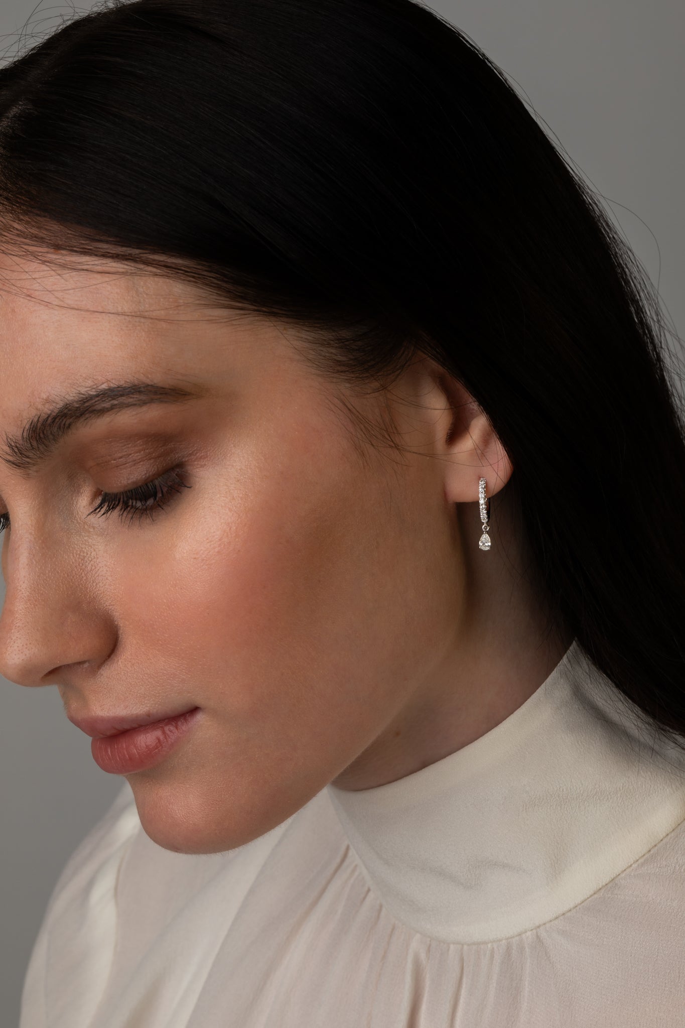 Profile view of a woman with dark hair wearing a white blouse and a silver drop earring featuring a small round diamond.
