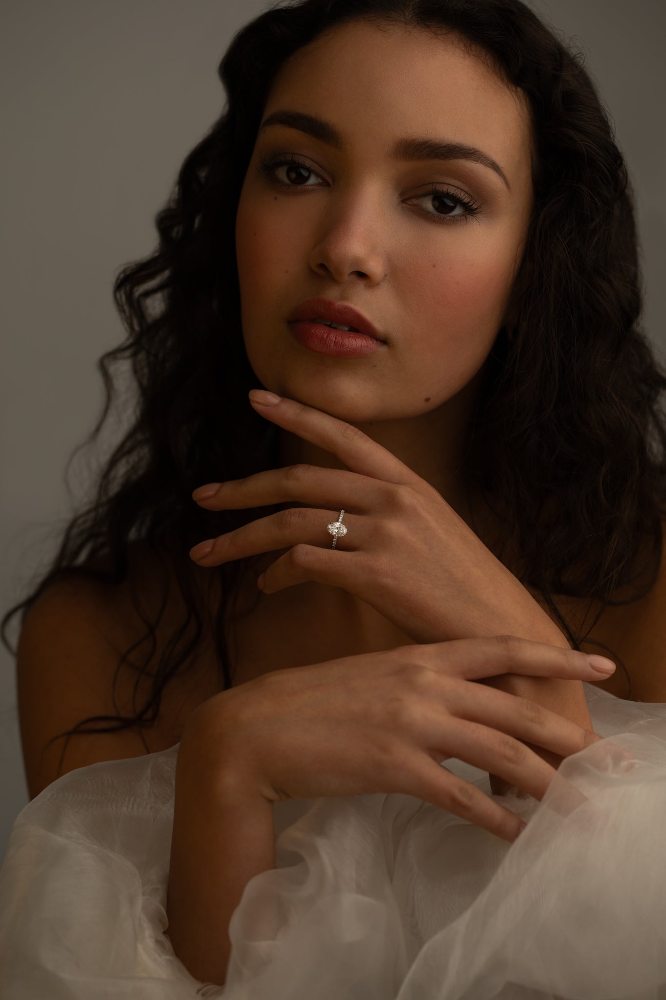 A close-up portrait of a woman with curly hair, wearing a white off-the-shoulder dress. She is gently touching her face with one hand, and a sparkling diamond ring is visible on her finger.