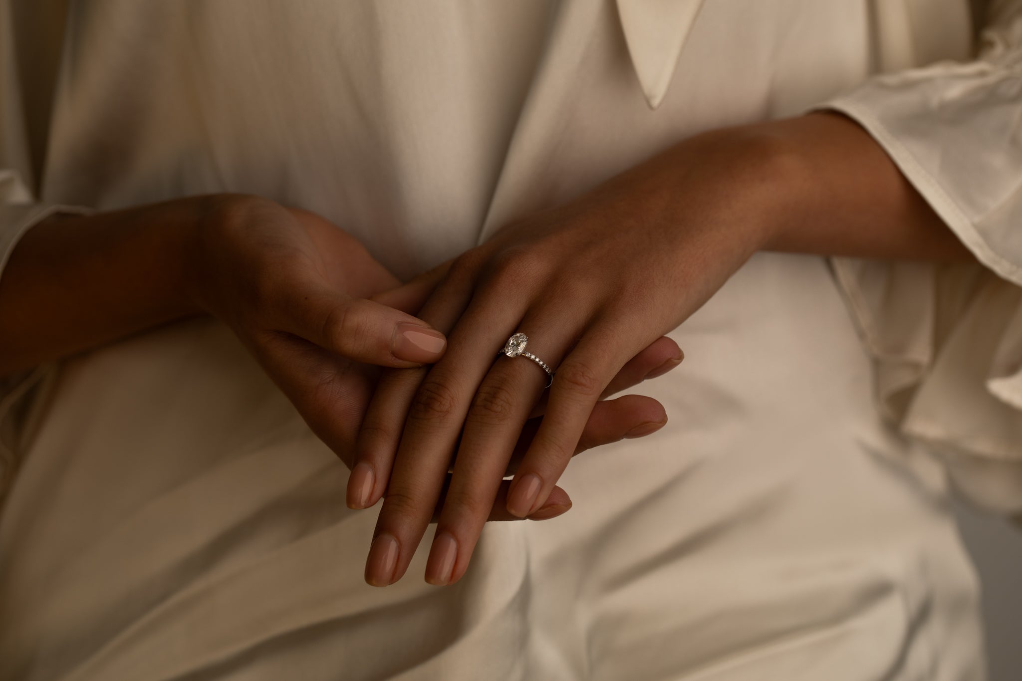 A close-up of a woman’s hands resting on her lap, showing a delicate diamond ring on her finger. She is wearing a white blouse with soft, flowing fabric.