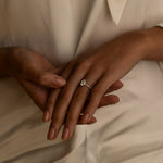 A close-up of a woman’s hands resting on her lap, showing a delicate diamond ring on her finger. She is wearing a white blouse with soft, flowing fabric.