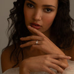 A woman with wavy hair is gently touching her face, wearing a delicate diamond ring on her finger. She gazes intently at the camera, surrounded by soft, flowing fabric.