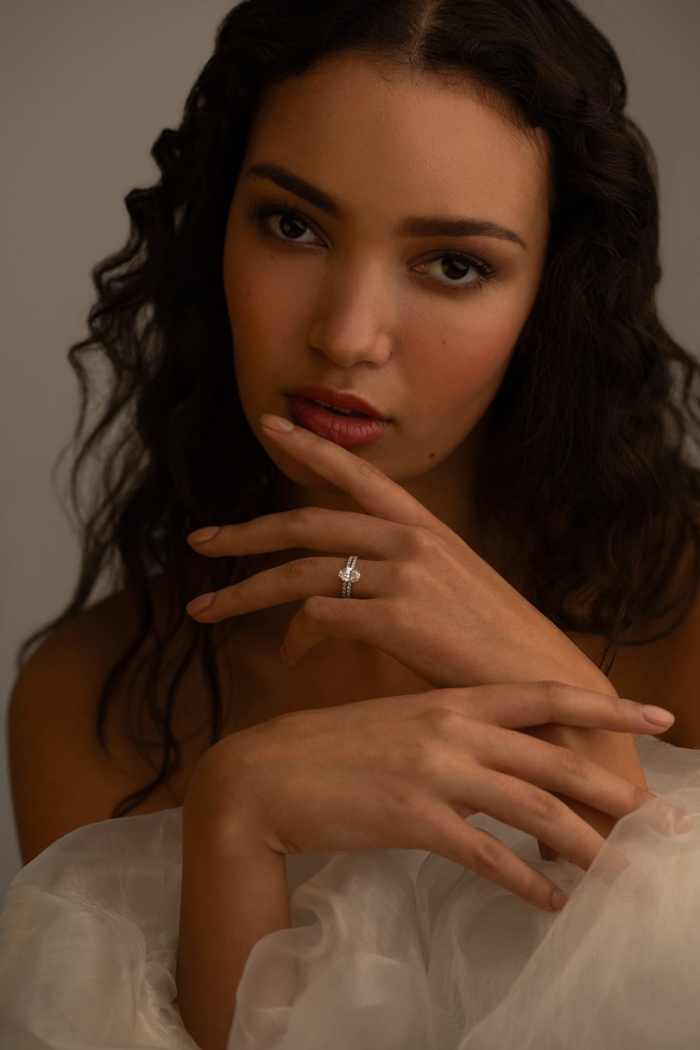 A woman with wavy hair is gently touching her face, wearing a delicate diamond ring on her finger. She gazes intently at the camera, surrounded by soft, flowing fabric.