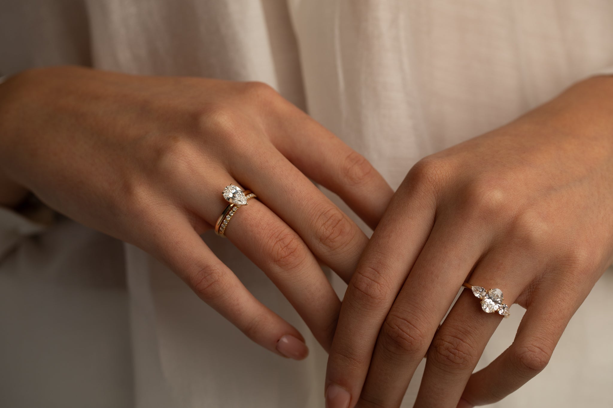 Two hands with rings featuring large center stones and diamond bands, set against a soft, neutral background, with one hand showcasing a pear-shaped diamond ring paired with a matching band, and the other hand displaying a three-stone ring.