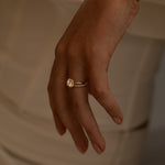 A close-up of a hand with a gold ring featuring a round center stone and a band adorned with smaller diamonds, resting against a soft, neutral-toned fabric.