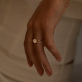 A close-up of a hand with a gold ring featuring a round center stone and a band adorned with smaller diamonds, resting against a soft, neutral-toned fabric.