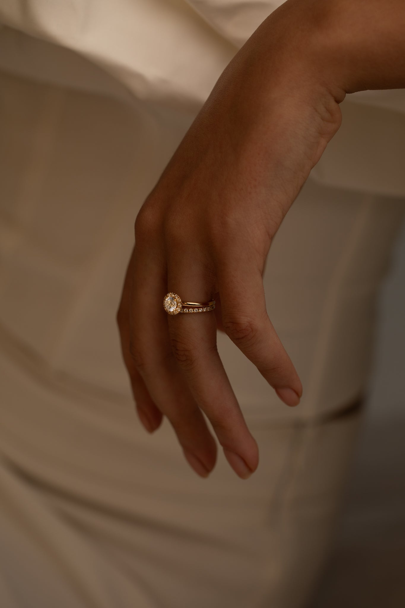 A close-up of a hand with a gold ring featuring a round center stone and a band adorned with smaller diamonds, resting against a soft, neutral-toned fabric.