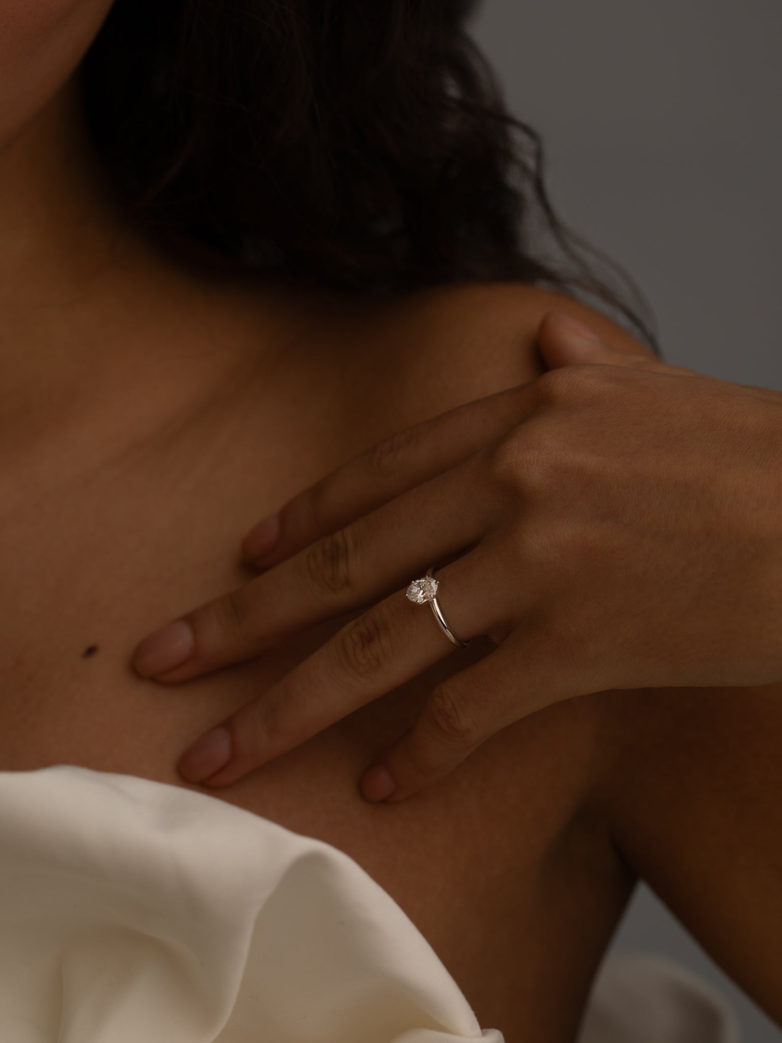 A close-up image of a woman's hand resting on her shoulder, showing a diamond ring on her finger. She is wearing a white off-the-shoulder top, and the background is neutral.