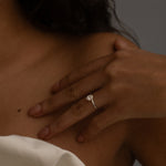 A close-up image of a woman's hand resting on her shoulder, showing a diamond ring on her finger. She is wearing a white off-the-shoulder top, and the background is neutral.