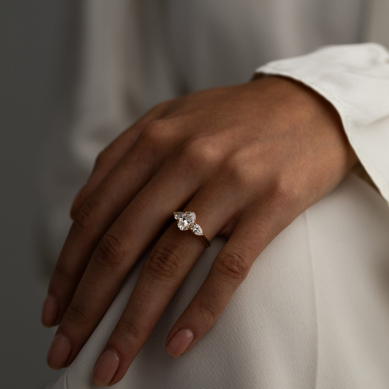 Hand resting on a knee, wearing a sparkling three-stone diamond ring with a pear-shaped center stone.