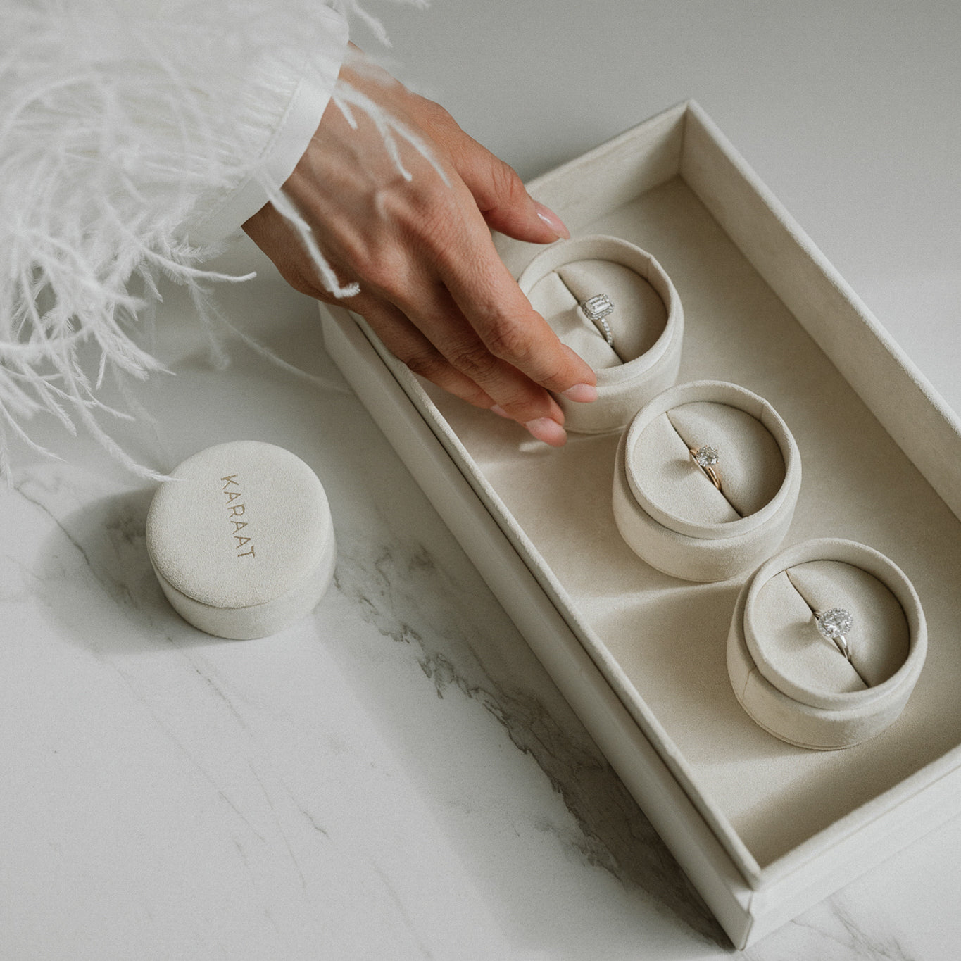A hand delicately selecting a ring from a cream-colored jewelry box containing three rings, placed on a marble surface.