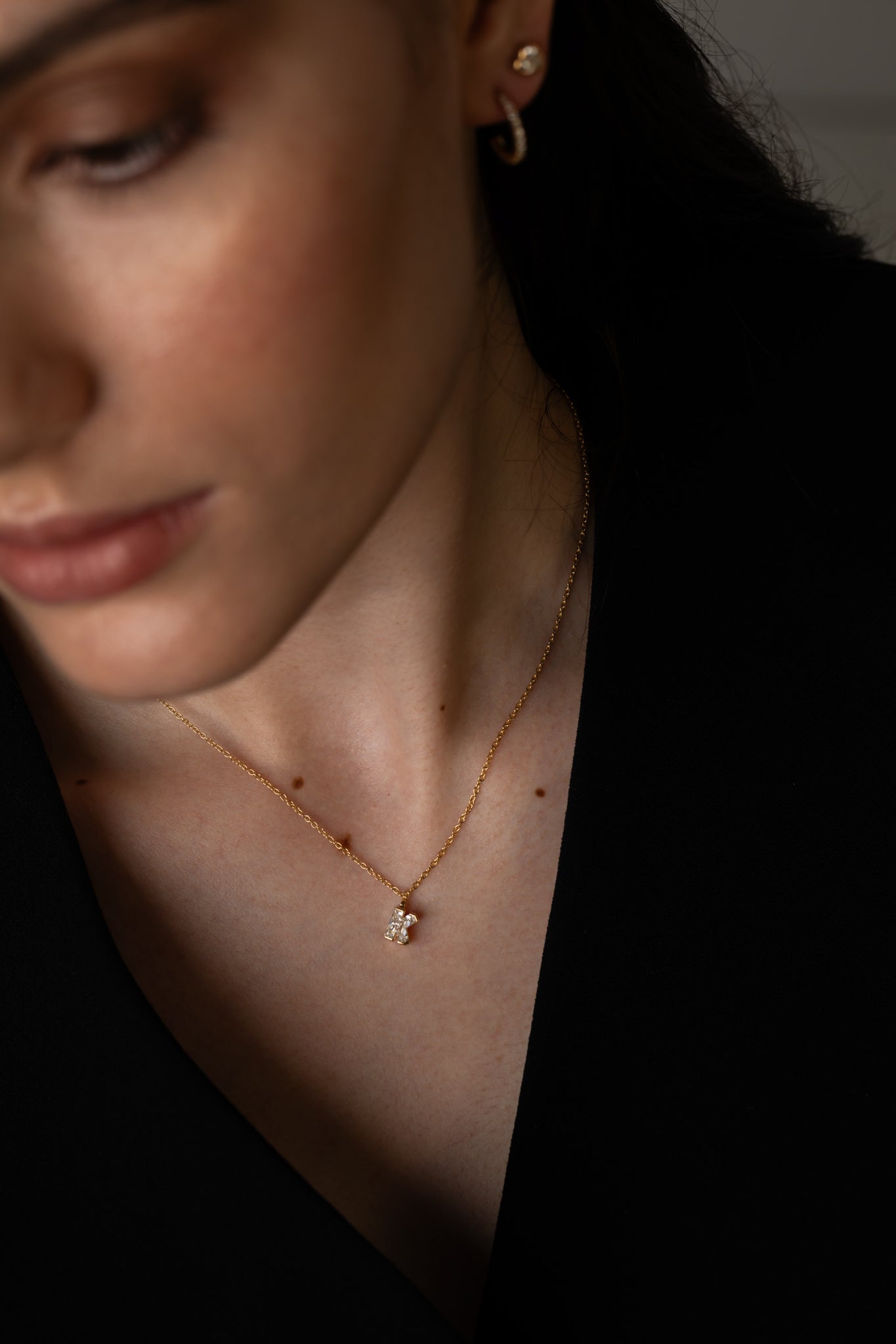 Close-up of a woman wearing a gold necklace with a small diamond pendant and matching earrings, dressed in black.