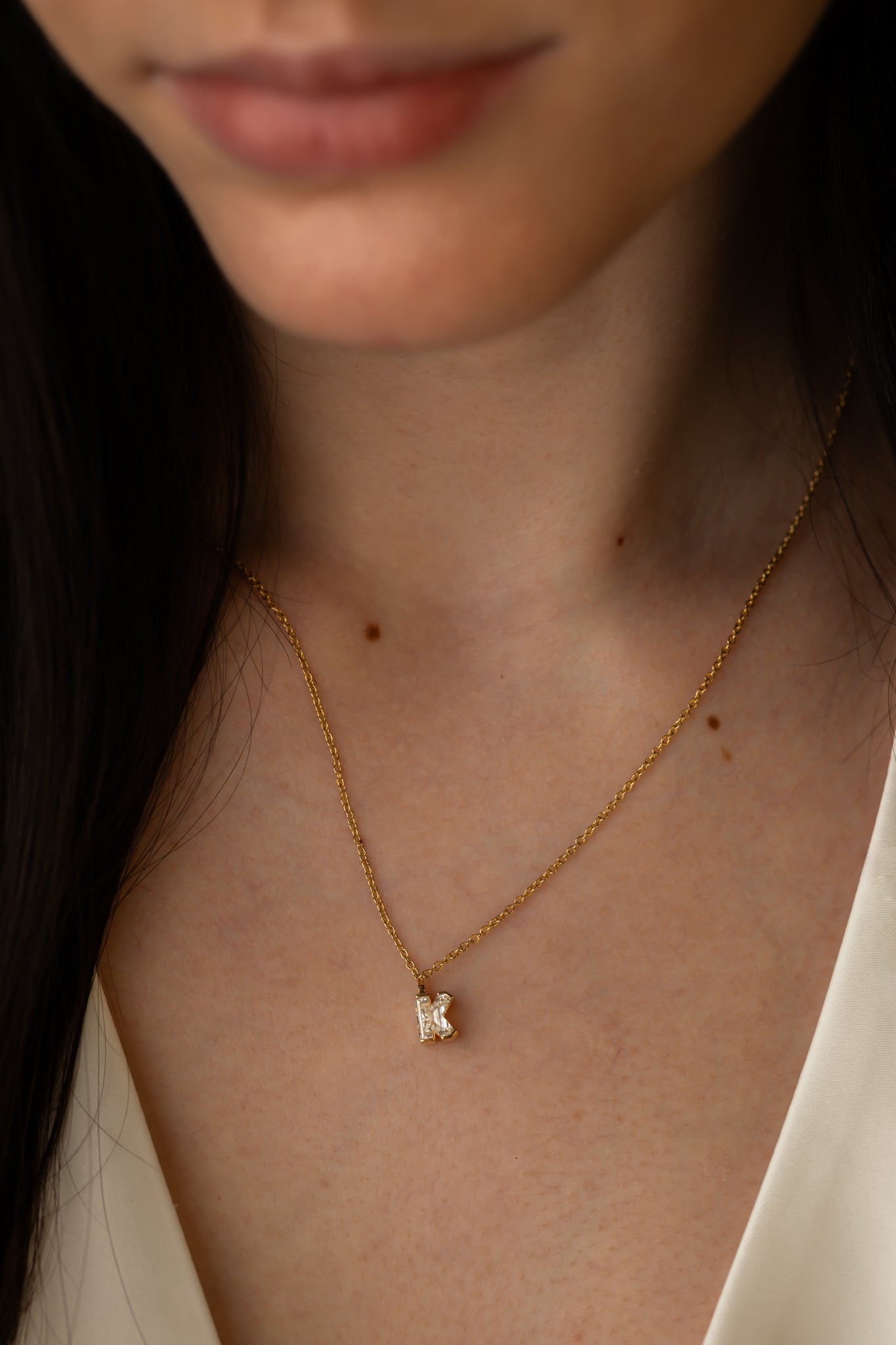 Close-up of a woman wearing a gold necklace with a diamond-studded "K" pendant, paired with a white top.