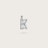Silver pendant in the shape of the letter "K," adorned with sparkling gemstones, displayed on a light beige background.
