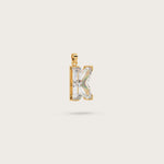 Gold pendant in the shape of the letter "K," adorned with sparkling gemstones, displayed on a light beige background.