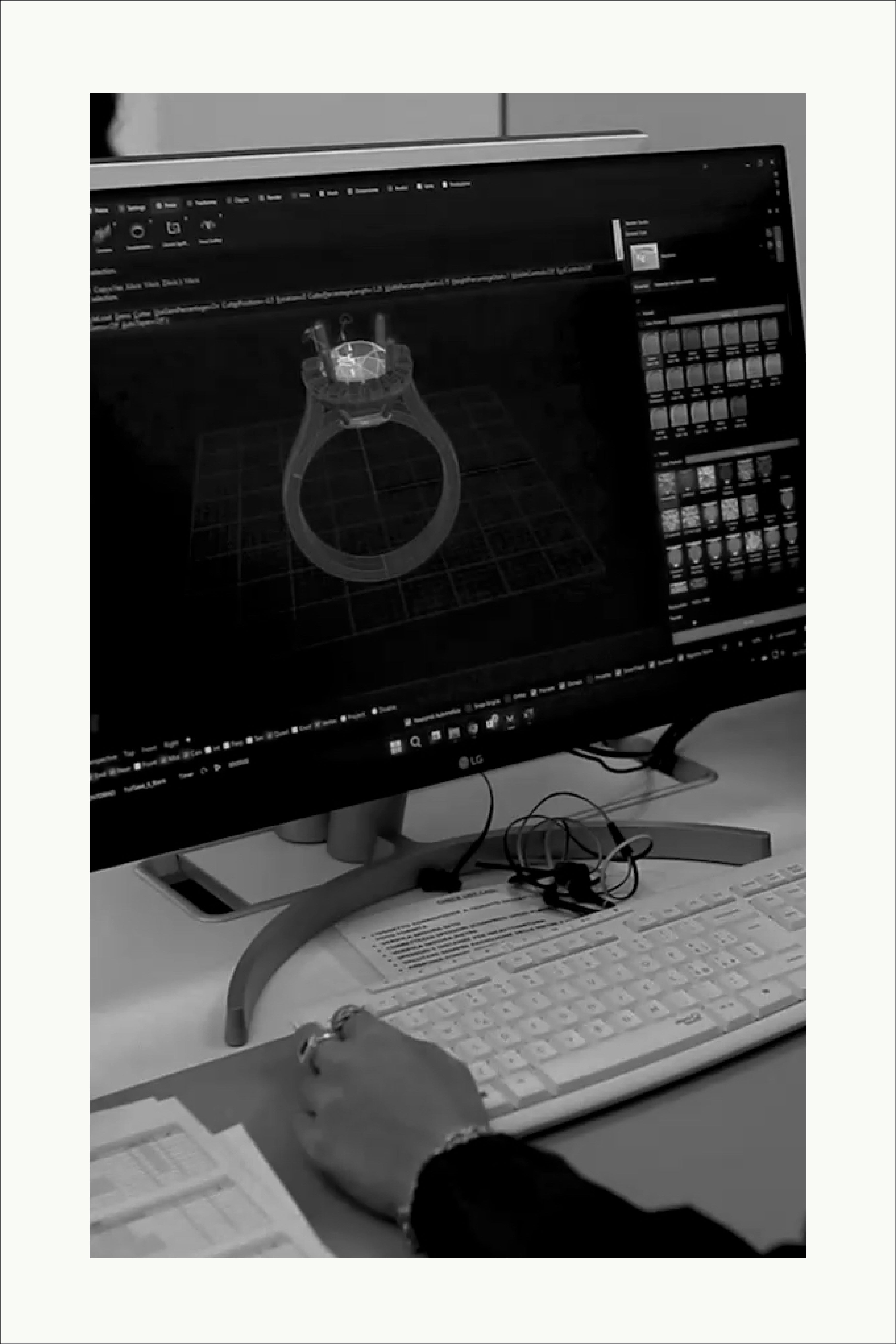 Designer working on a 3D digital model of a ring displayed on a computer monitor, with a keyboard and notes on the desk.