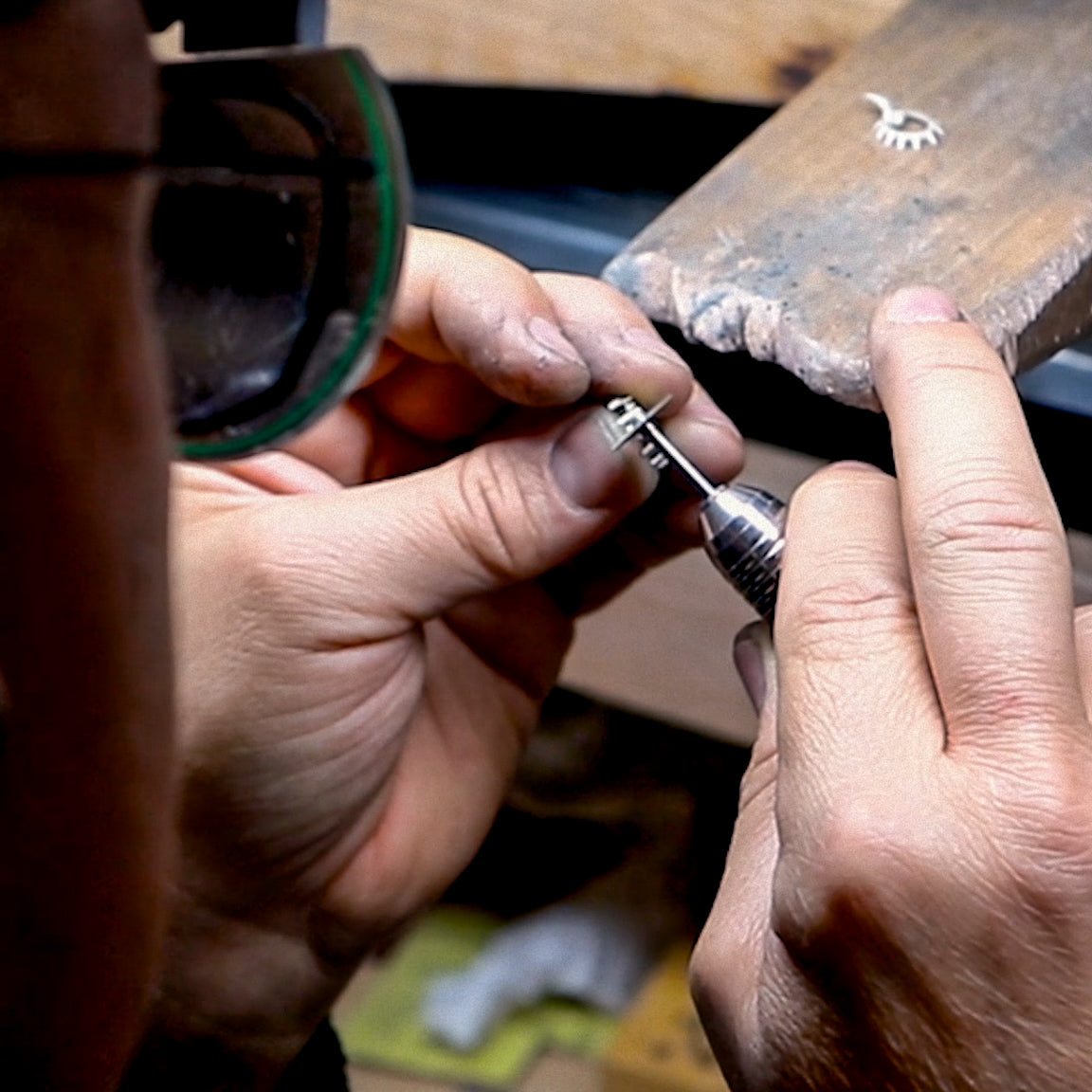 A jewelry artisan wearing magnifying glasses, carefully working on a silver piece using a precision tool at a wooden workstation.






