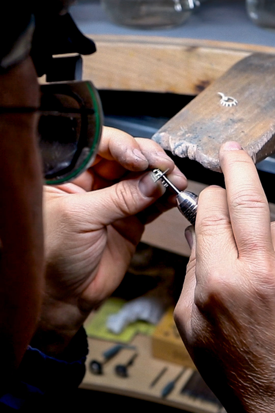 A jewelry artisan wearing magnifying glasses, carefully working on a silver piece using a precision tool at a wooden workstation.






