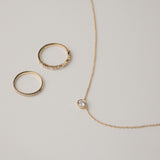 A minimalist flat-lay product photograph featuring a round bezel-set diamond pendant necklace displayed beside two matching delicate gold pavé diamond stacking rings on a light grey background.