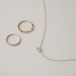 A minimalist flat-lay product photograph featuring a round white gold bezel-set diamond pendant necklace displayed beside two matching delicate gold pavé diamond stacking rings on a light grey background.