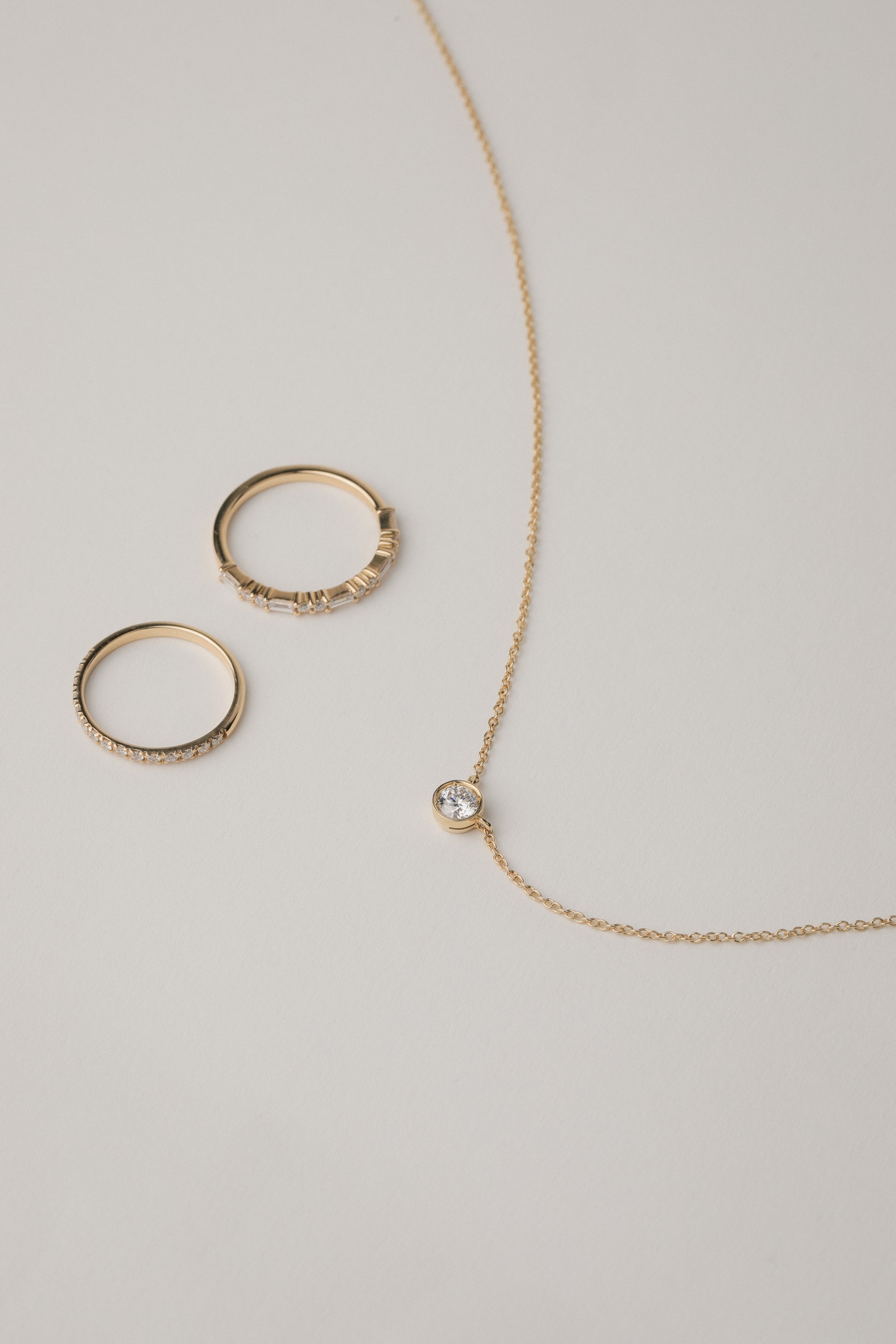 A minimalist flat-lay product photograph featuring a round white gold bezel-set diamond pendant necklace displayed beside two matching delicate gold pavé diamond stacking rings on a light grey background.