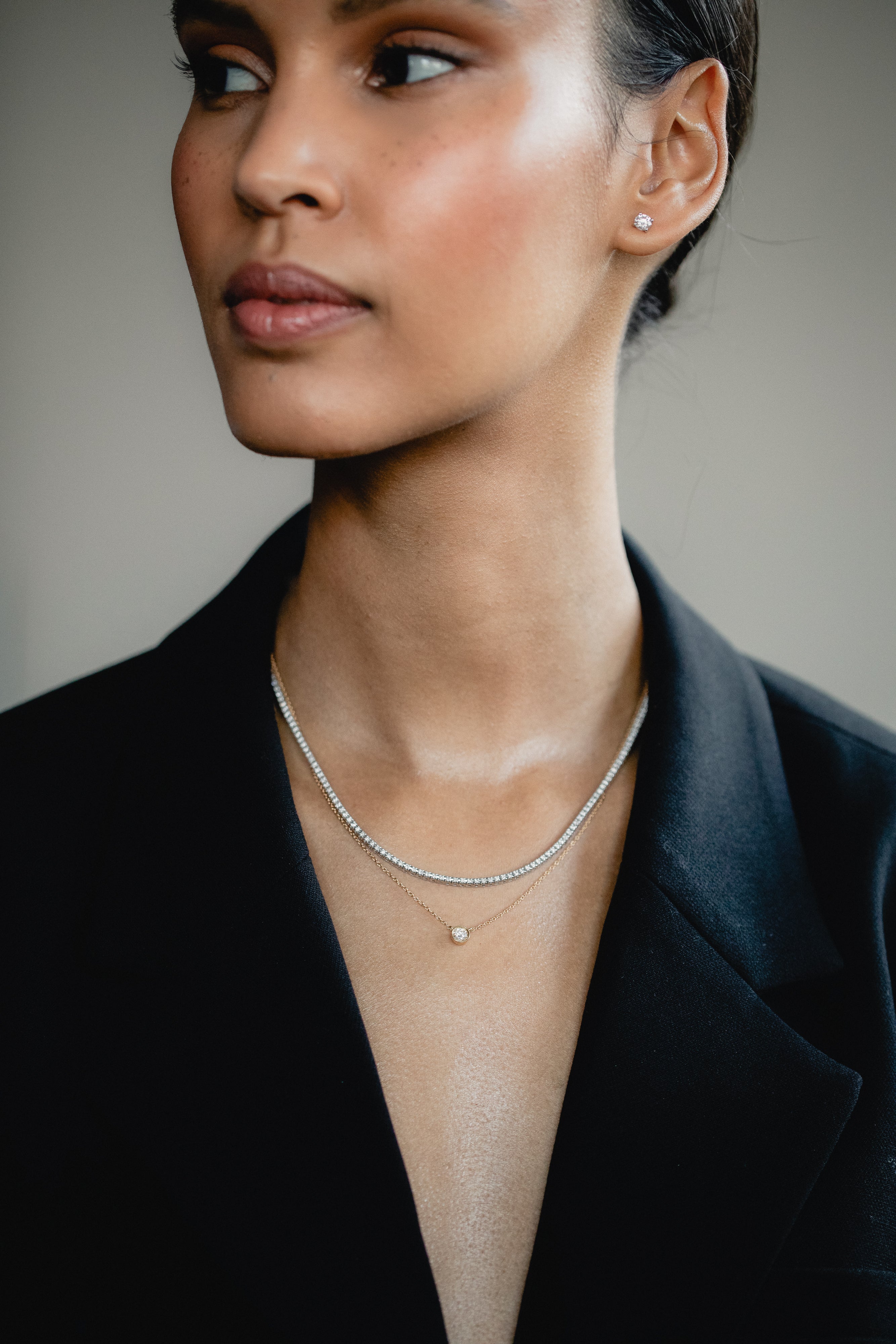 An elegant editorial lifestyle portrait of a model wearing a blazer, showcasing a luxury layering combination with a white gold bezel-set diamond necklace layered beneath a shimmering rose gold diamond tennis necklace.