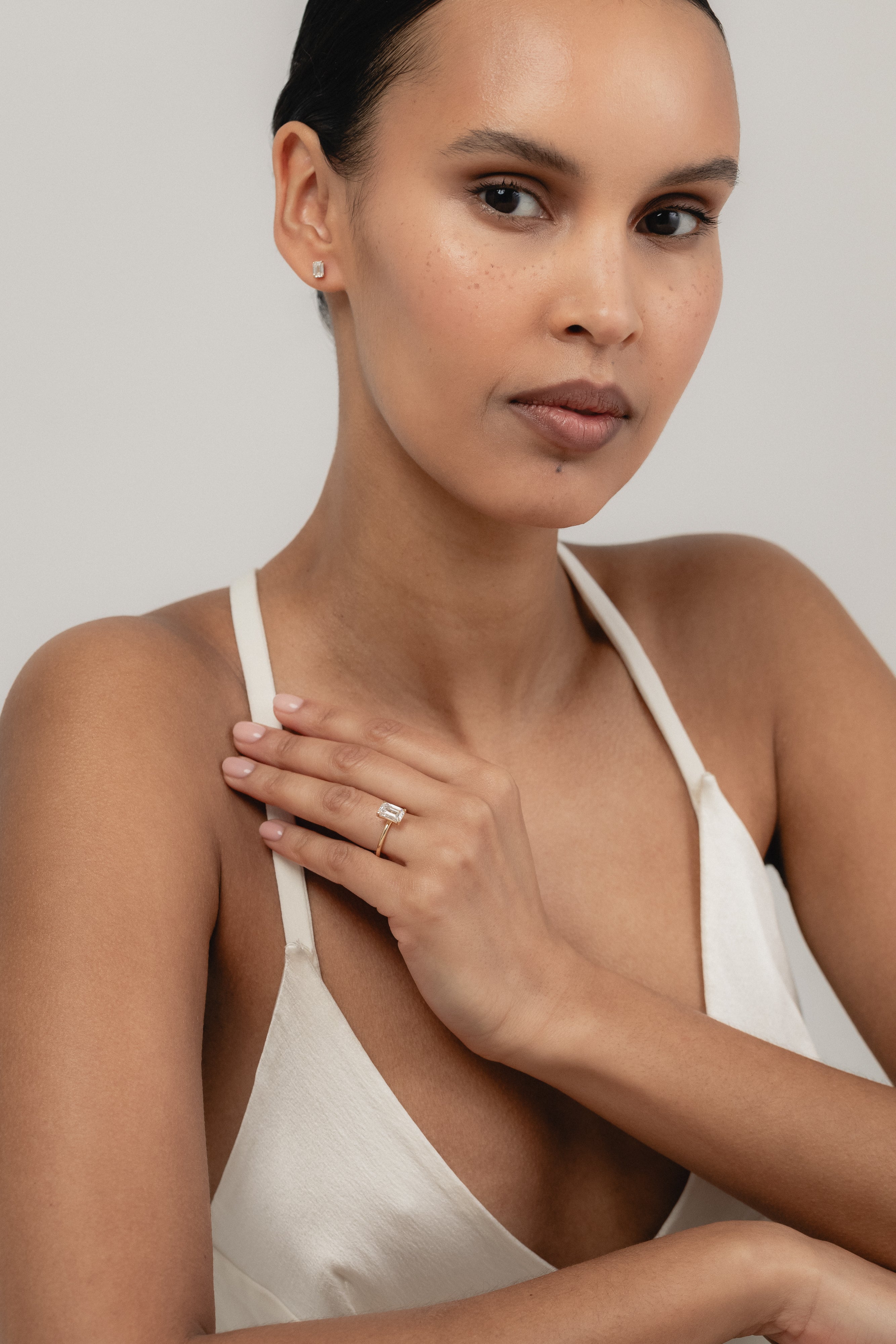 An editorial portrait of a model wearing a white strap top, elegantly displaying a yellow gold KARAAT emerald-shaped diamond solitaire ring stacked with a gold band on her finger.