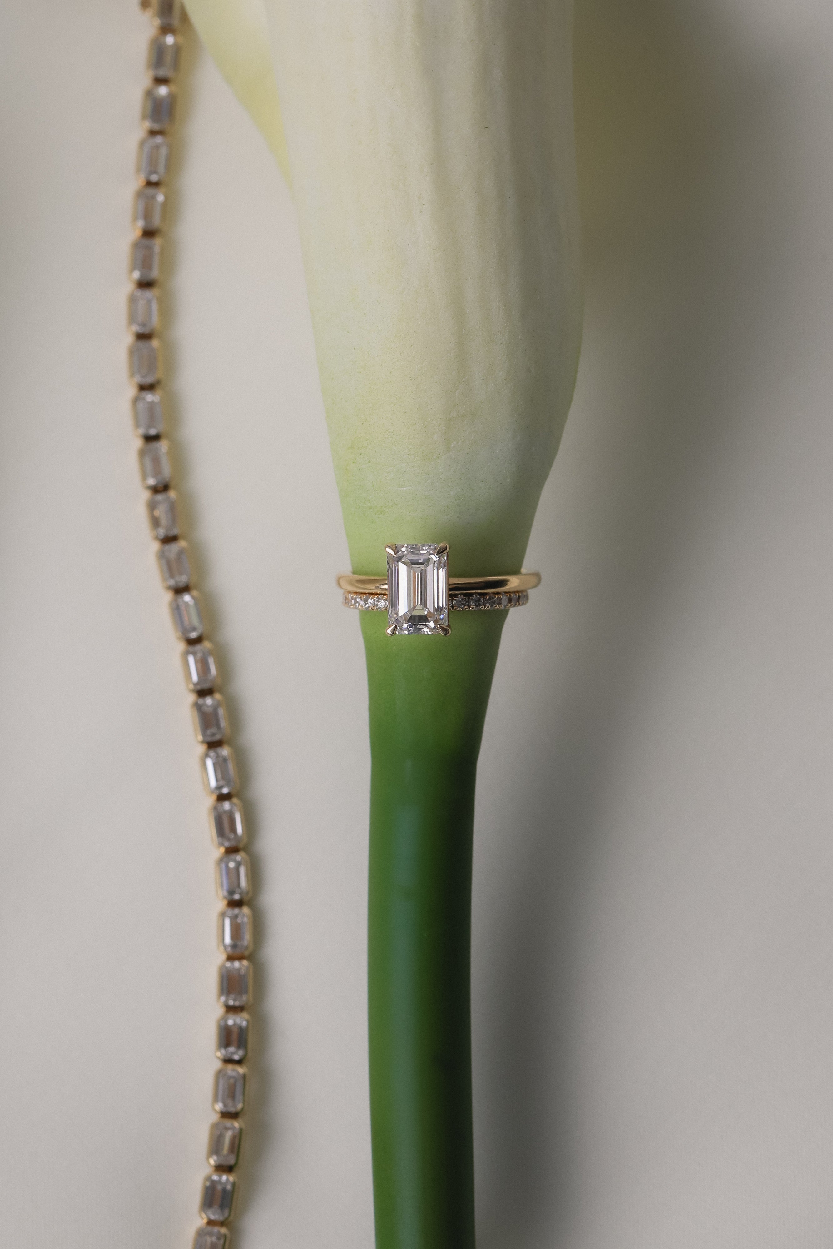 An artistic still-life photograph of a yellow gold KARAAT emerald-cut diamond engagement ring slipped onto the stem of a white calla lily, placed next to a shimmering diamond tennis bracelet.