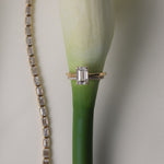 An artistic still-life photograph of a white gold KARAAT emerald-cut diamond engagement ring slipped onto the stem of a white calla lily, placed next to a shimmering diamond tennis bracelet.