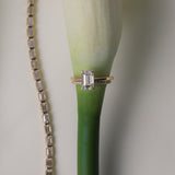 An artistic still-life photograph of a white gold KARAAT emerald-cut diamond engagement ring slipped onto the stem of a white calla lily, placed next to a shimmering diamond tennis bracelet.