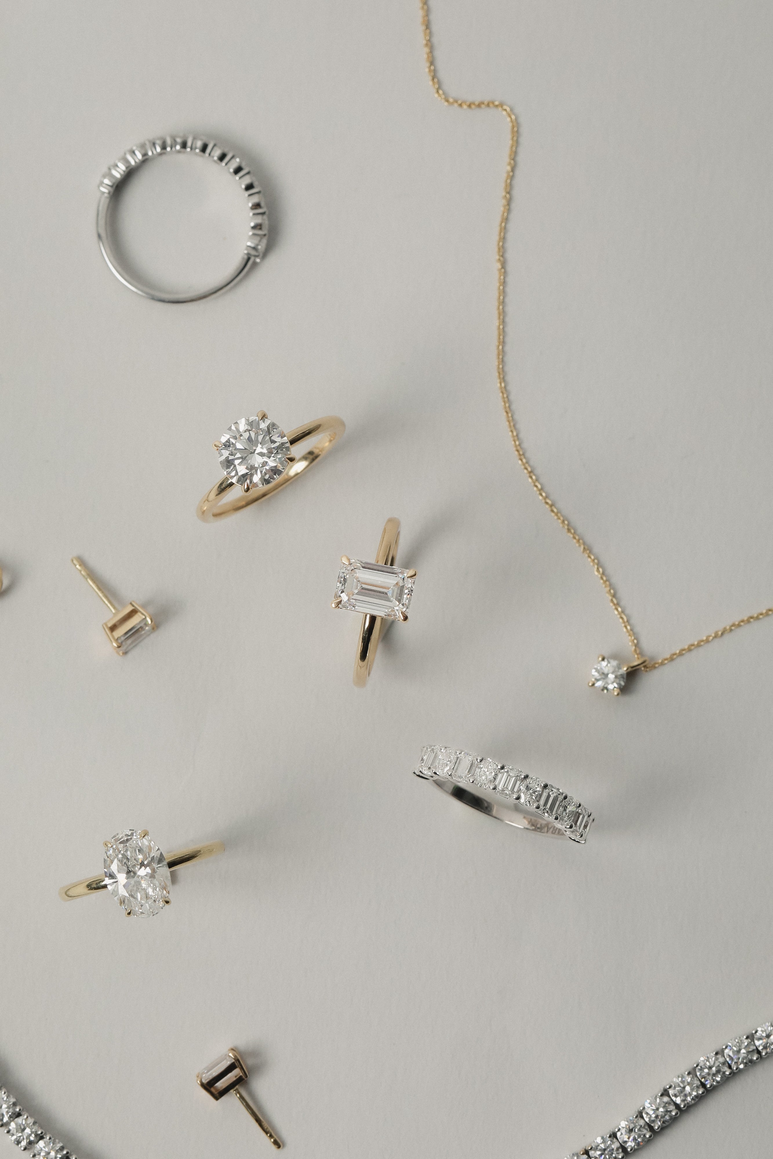 A luxurious flat-lay arrangement of KARAAT fine jewelry on a neutral grey background, featuring a yellow gold emerald-cut engagement ring, an oval solitaire, a round diamond ring, a tennis bracelet, and a delicate diamond pendant necklace.